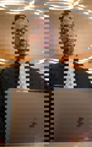 Faruk draagt zijn werkkleding (advocaat) en staat achter een microfoon. Zijn laptop staat voor hem. Hij is aan het praten. Faruk draagt zijn werkkleding (advocaat) en staat achter een microfoon. Zijn laptop staat voor hem. Hij is aan het praten.