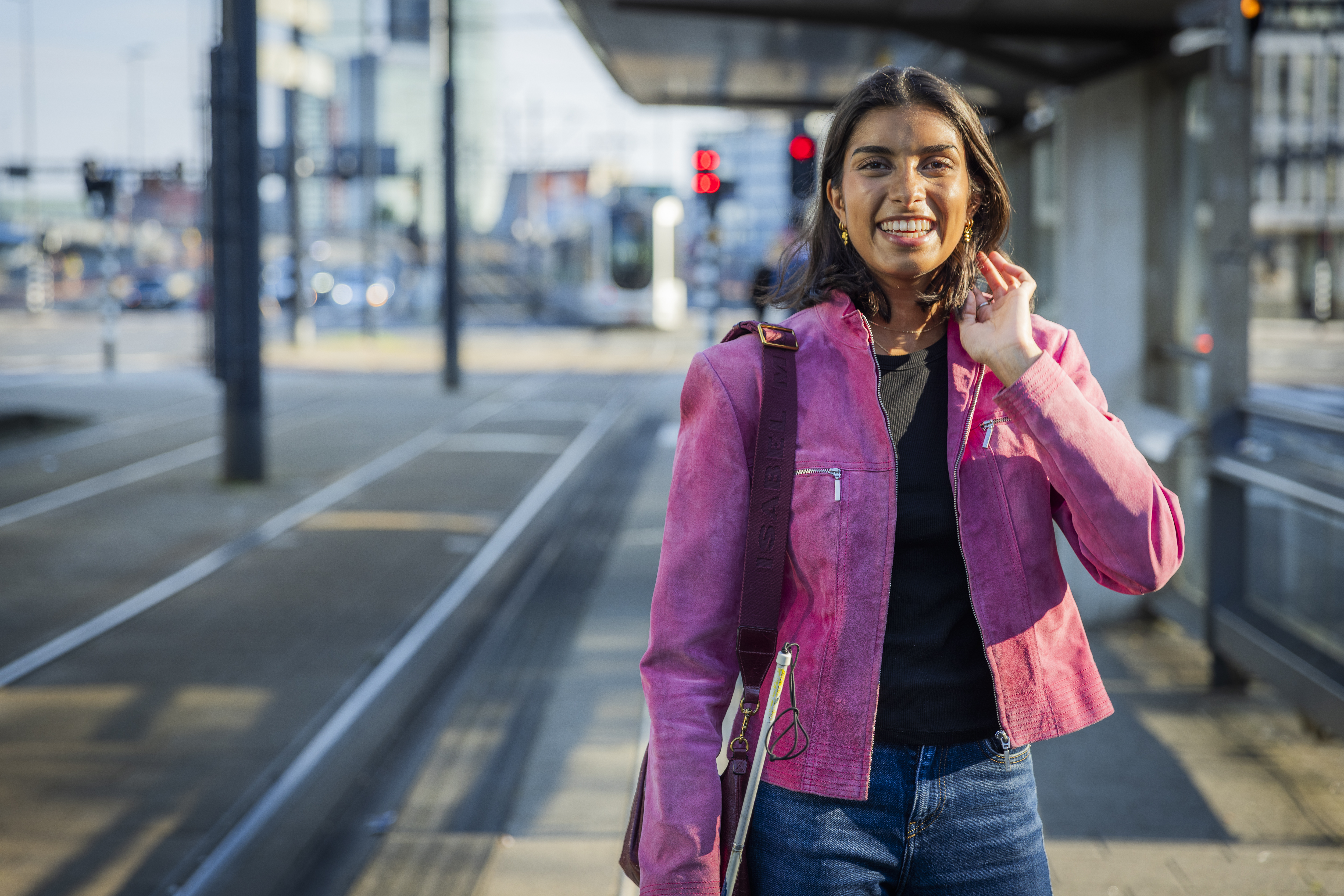Visio-ambassadeur Bodhi Sripal staat bij een tramhalte met haar taststok. De foto laat zien hoe Bodhi als slechtziende jongvolwassen vrouw zelfstandig gebruikmaakt van het openbaar vervoer.