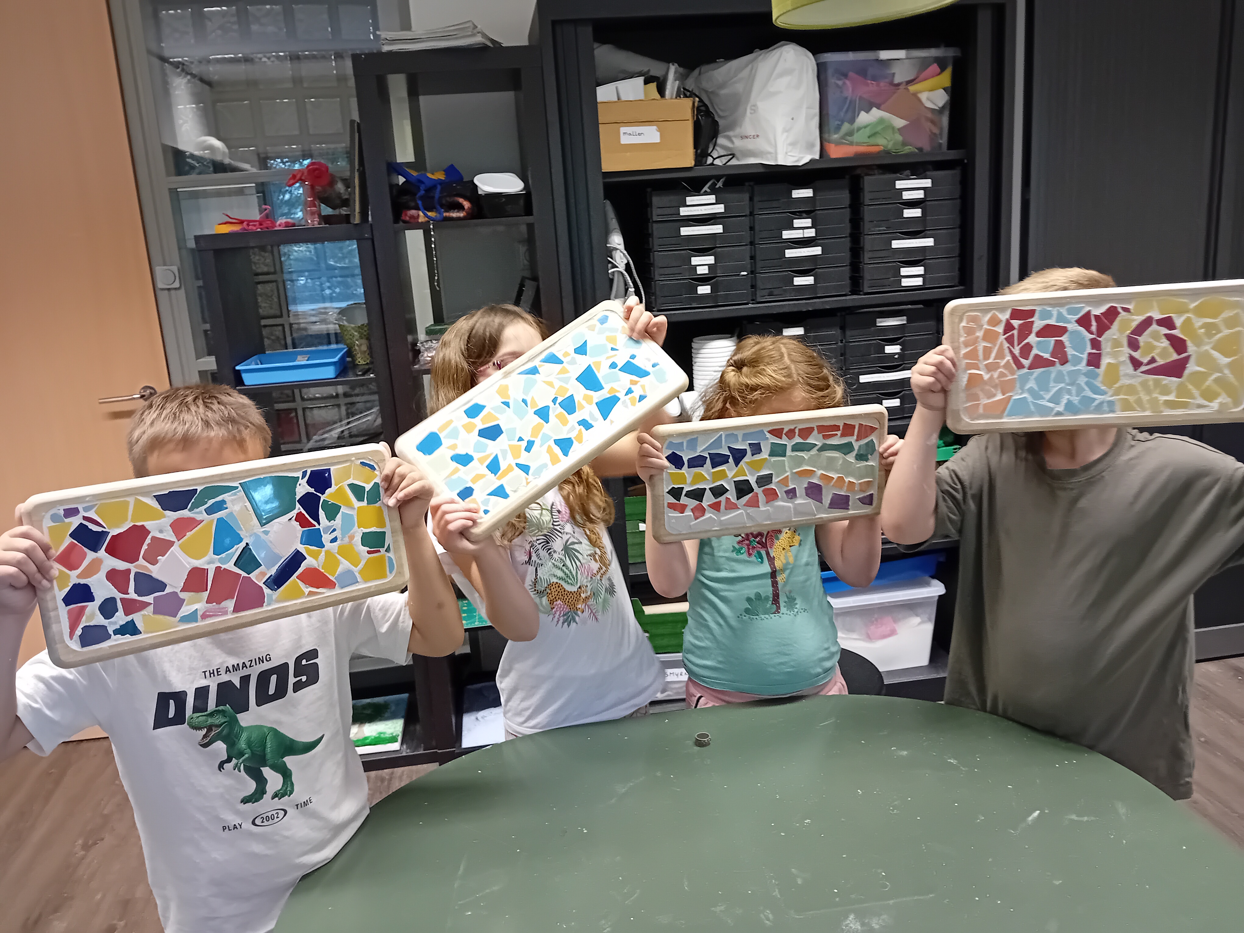 Vier kinderen houden elk een rechthoekig plateau omhoog, versierd met kleurrijke stukjes keramiek in mozaïekpatronen, gemaakt tijdens een knutselactiviteit bij Visio met kasten en materialen op de achtergrond.