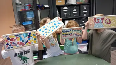 Vier kinderen houden elk een rechthoekig plateau omhoog, versierd met kleurrijke stukjes keramiek in mozaïekpatronen, gemaakt tijdens een knutselactiviteit bij Visio met kasten en materialen op de achtergrond. Vier kinderen houden elk een rechthoekig plateau omhoog, versierd met kleurrijke stukjes keramiek in mozaïekpatronen, gemaakt tijdens een knutselactiviteit bij Visio met kasten en materialen op de achtergrond.