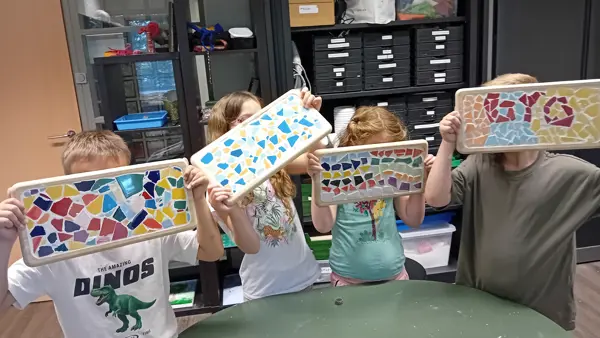 Vier kinderen houden elk een rechthoekig plateau omhoog, versierd met kleurrijke stukjes keramiek in mozaïekpatronen, gemaakt tijdens een knutselactiviteit bij Visio met kasten en materialen op de achtergrond. Vier kinderen houden elk een rechthoekig plateau omhoog, versierd met kleurrijke stukjes keramiek in mozaïekpatronen, gemaakt tijdens een knutselactiviteit bij Visio met kasten en materialen op de achtergrond.