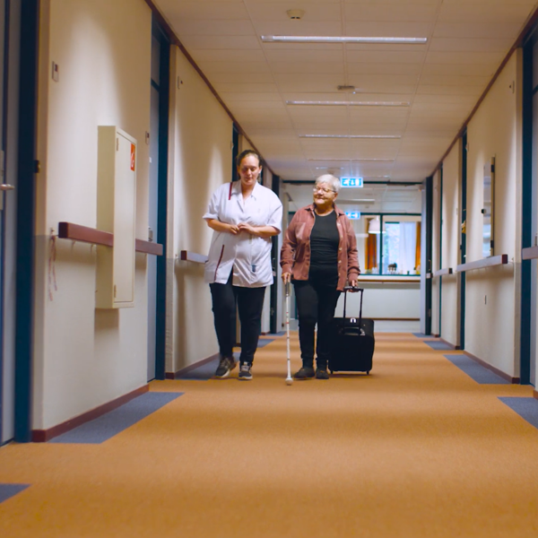 Cliënt met een taststok en een medewerker lopen samen door een hal van De Vlasborch Cliënt met een taststok en een medewerker lopen samen door een hal van De Vlasborch