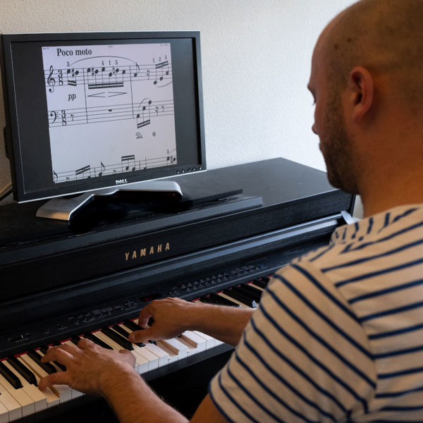 Cliënt van Visio Het Loo Erf speelt piano waarbij de bladmuziek vergroot is op een beeldscherm Cliënt van Visio Het Loo Erf speelt piano waarbij de bladmuziek vergroot is op een beeldscherm