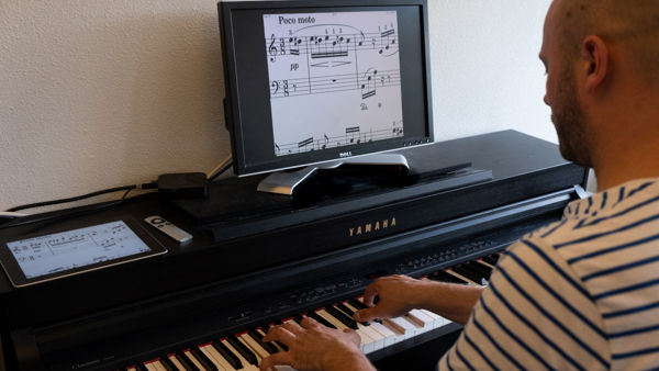 Cliënt van Visio Het Loo Erf speelt piano waarbij de bladmuziek vergroot is op een beeldscherm Cliënt van Visio Het Loo Erf speelt piano waarbij de bladmuziek vergroot is op een beeldscherm