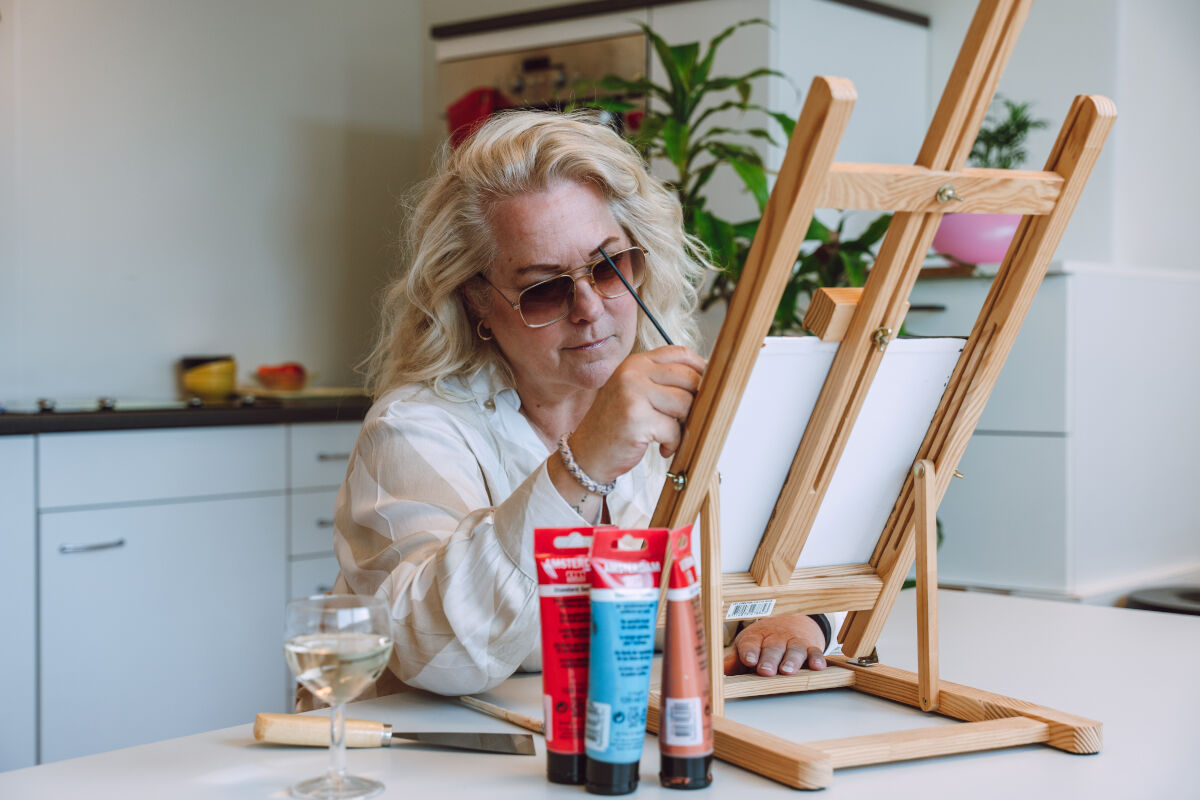 Een blonde vrouw zit aan een tafel. Ze is aan het schilderen. 