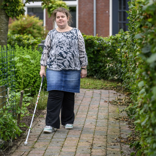 Dame met taststok loopt buiten op een stoepje bij Visio Wonen & Dagbesteding Stramproy Dame met taststok loopt buiten op een stoepje bij Visio Wonen & Dagbesteding Stramproy
