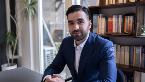 Foto van Faruk, hij zit aan een bureau in een kantoor met een blauw pak aan. Foto van Faruk, hij zit aan een bureau in een kantoor met een blauw pak aan.