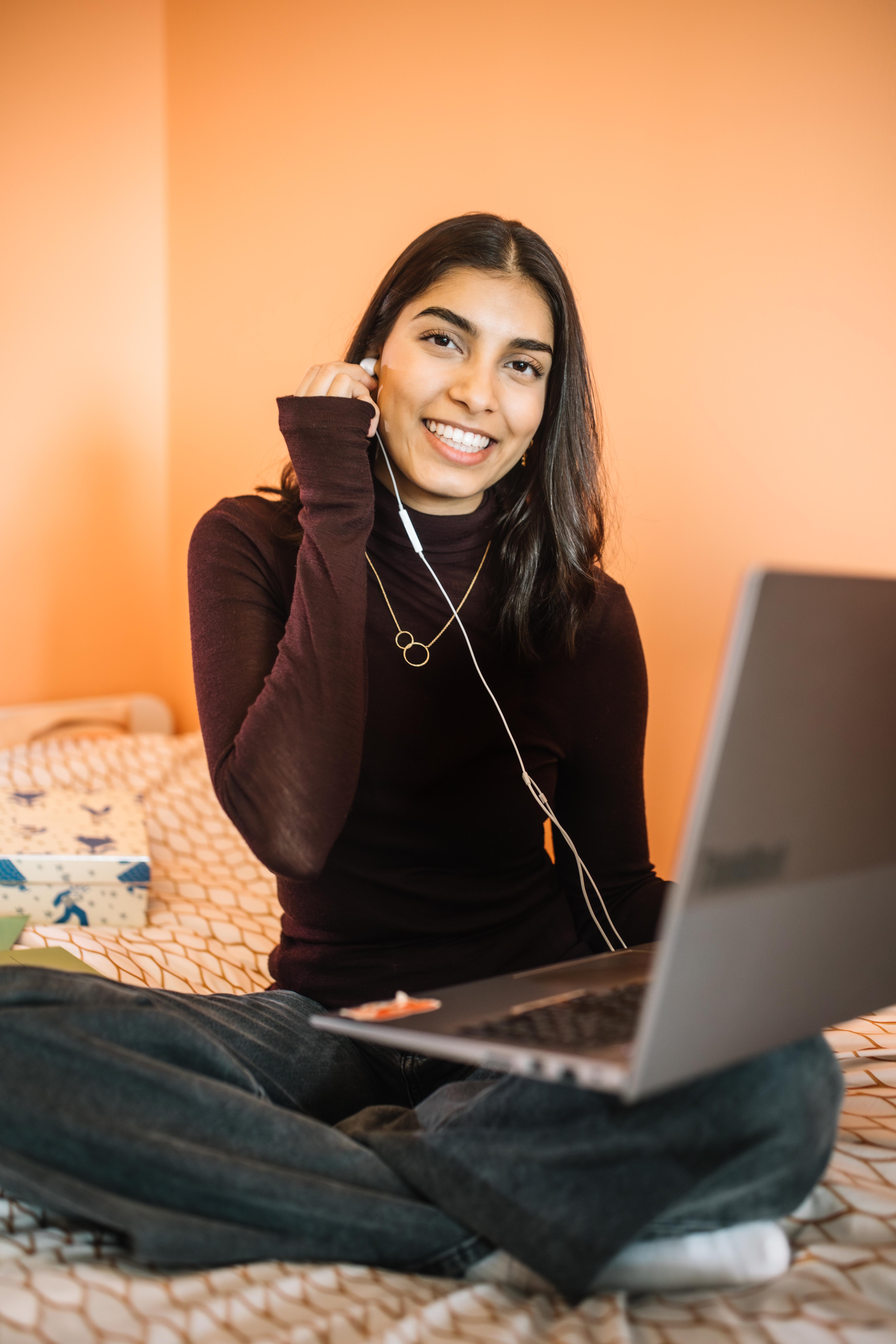 Bodhi zit op haar bed met haar laptop. Ze doet een oordopje, aangesloten aan haar laptop, in haar oor. 