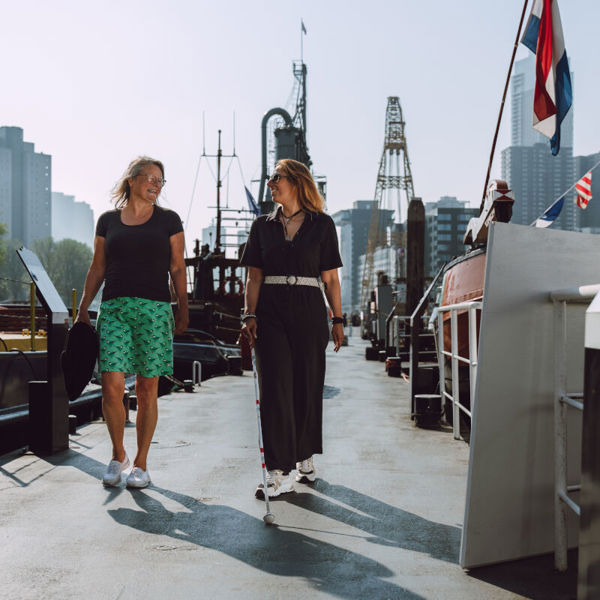 Twee vrouwen lopen pratend op straat, waarvan een vrouw een taststok in haar handen heeft Twee vrouwen lopen pratend op straat, waarvan een vrouw een taststok in haar handen heeft