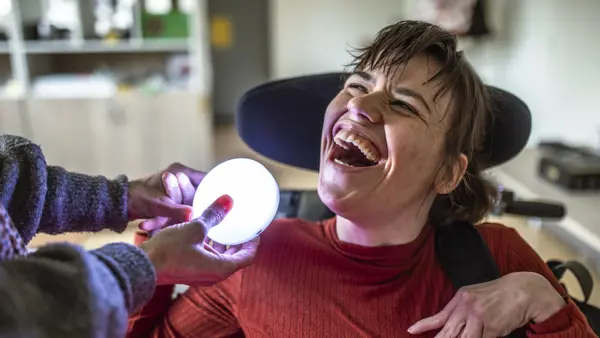 Een vrouw in een rolstoel heeft een grote glimlach op haar gezicht. Een begeleider houdt een lichtgevend object vast voor haar. Een vrouw in een rolstoel heeft een grote glimlach op haar gezicht. Een begeleider houdt een lichtgevend object vast voor haar.