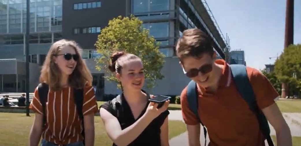 Drie leerlingen lopen samen. Het middelste meisje laat de jongen aan haar linkerkant iets horen met haar telefoon. 
