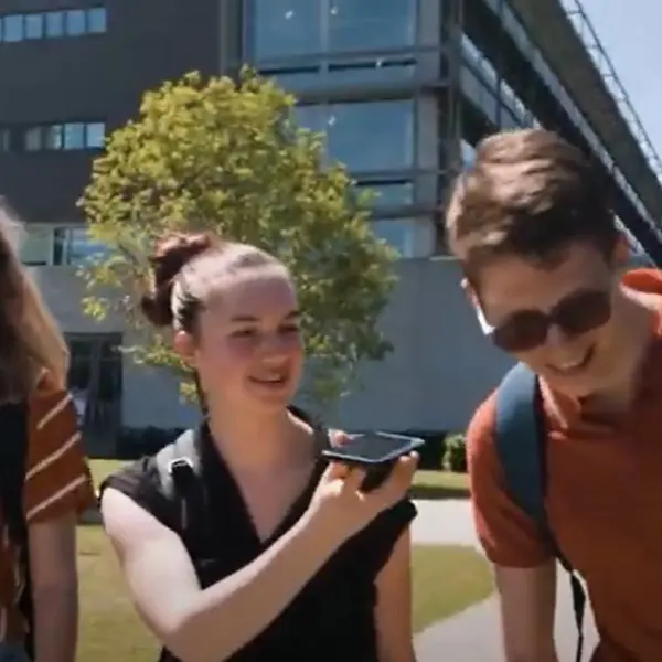 Drie leerlingen lopen samen. Het middelste meisje laat de jongen aan haar linkerkant iets horen met haar telefoon. Drie leerlingen lopen samen. Het middelste meisje laat de jongen aan haar linkerkant iets horen met haar telefoon.