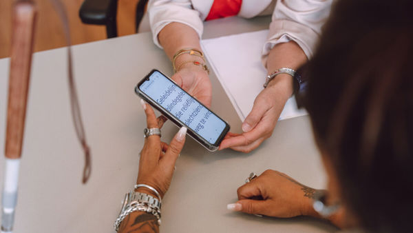 Twee paar handen houden een telefoon vast. Er staat vergrote tekst in beeld. Twee paar handen houden een telefoon vast. Er staat vergrote tekst in beeld.