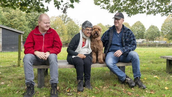 Drie cliënten van Visio zitten samen buiten op een houten bankje, één van hen knuffelt een hond Drie cliënten van Visio zitten samen buiten op een houten bankje, één van hen knuffelt een hond