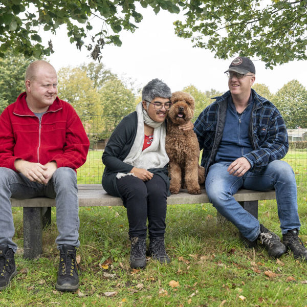 Drie cliënten van Visio zitten samen buiten op een houten bankje, één van hen knuffelt een hond Drie cliënten van Visio zitten samen buiten op een houten bankje, één van hen knuffelt een hond