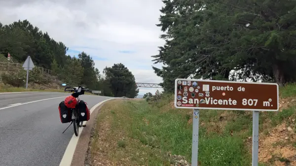 Fiets met bepakking staat geparkeerd aan de rand van een autoweg, daarbij een bord met "Puerto de San Vicente 807 m." Fiets met bepakking staat geparkeerd aan de rand van een autoweg, daarbij een bord met "Puerto de San Vicente 807 m."