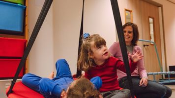 Twee kinderen spelen binnen in een schommelmand met een begeleider erbij Twee kinderen spelen binnen in een schommelmand met een begeleider erbij