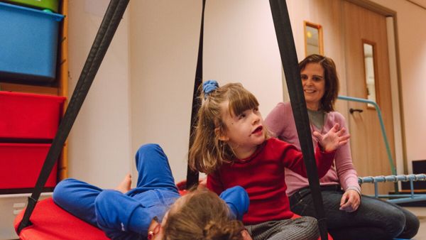 Twee kinderen spelen binnen in een schommelmand met een begeleider erbij Twee kinderen spelen binnen in een schommelmand met een begeleider erbij