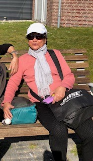 Maureen zit op een bankje in de zon. Ze draagt een pet en een zonnebril. 