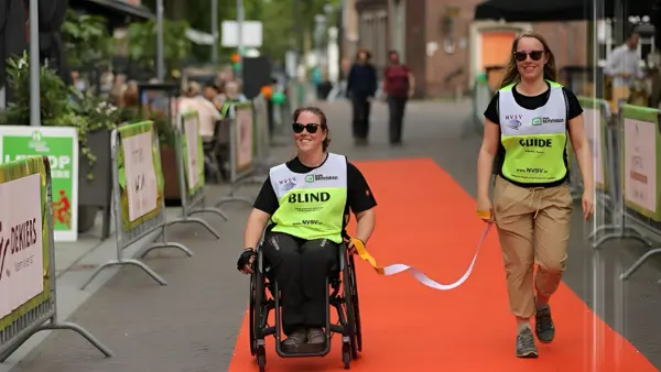 Desiree Vranken samen met haar wandelbuddy op een rode loper tijdens een sportevenement, zij zijn verbonden door een koord dat zij beiden vasthouden Desiree Vranken samen met haar wandelbuddy op een rode loper tijdens een sportevenement, zij zijn verbonden door een koord dat zij beiden vasthouden