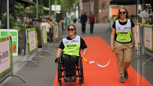 Desiree Vranken samen met haar wandelbuddy op een rode loper tijdens een sportevenement, zij zijn verbonden door een koord dat zij beiden vasthouden Desiree Vranken samen met haar wandelbuddy op een rode loper tijdens een sportevenement, zij zijn verbonden door een koord dat zij beiden vasthouden