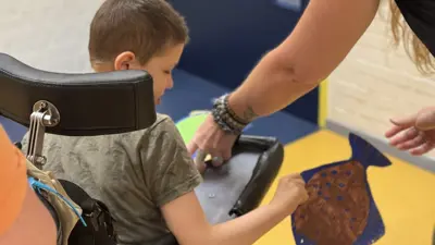 Begeleider helpt een kind in een rolstoel terwijl het kind een kleurrijke vis vasthoudt; in een gang met gele vloer en blauwe deur. Begeleider helpt een kind in een rolstoel terwijl het kind een kleurrijke vis vasthoudt; in een gang met gele vloer en blauwe deur.