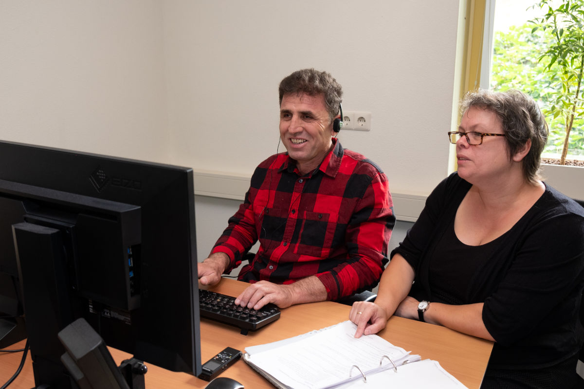 Cliënt en trainer van Visio Het Loo Erf zitten tijdens een ICT-training naast elkaar aan een bureau waarbij de cliënt oefent met het gebruik van de computer