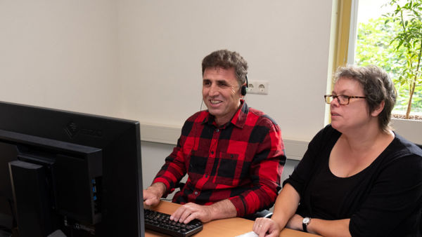 Cliënt en trainer van Visio Het Loo Erf zitten tijdens een ICT-training naast elkaar aan een bureau waarbij de cliënt oefent met het gebruik van de computer Cliënt en trainer van Visio Het Loo Erf zitten tijdens een ICT-training naast elkaar aan een bureau waarbij de cliënt oefent met het gebruik van de computer