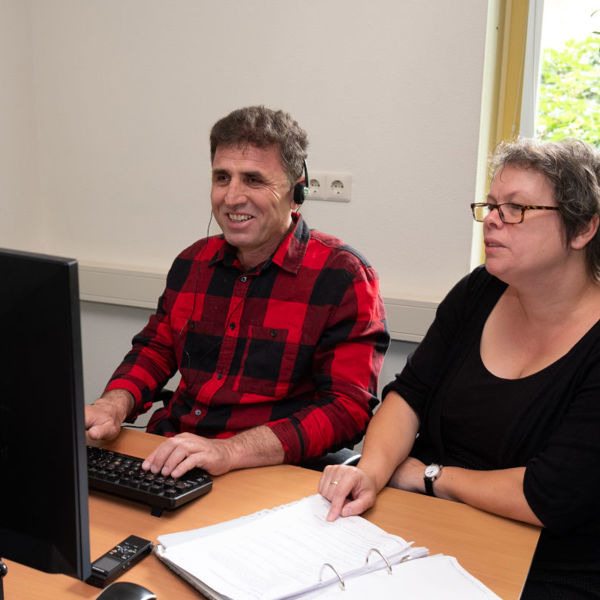 Cliënt en trainer van Visio Het Loo Erf zitten tijdens een ICT-training naast elkaar aan een bureau waarbij de cliënt oefent met het gebruik van de computer Cliënt en trainer van Visio Het Loo Erf zitten tijdens een ICT-training naast elkaar aan een bureau waarbij de cliënt oefent met het gebruik van de computer