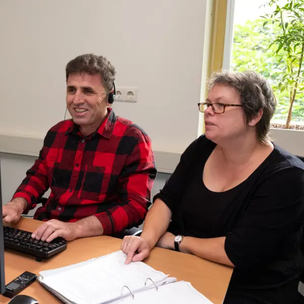 Cliënt en trainer van Visio Het Loo Erf zitten tijdens een ICT-training naast elkaar aan een bureau waarbij de cliënt oefent met het gebruik van de computer Cliënt en trainer van Visio Het Loo Erf zitten tijdens een ICT-training naast elkaar aan een bureau waarbij de cliënt oefent met het gebruik van de computer