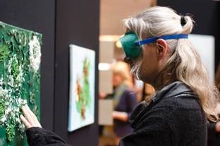 Blonde vrouw met groene verduisterde bril voelt aan een kunstwerk aan de muur.